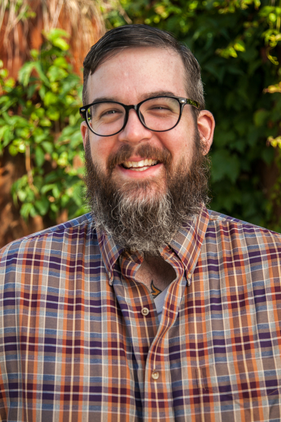 A smiling bearded white man in a plaid shirt with a green deciduous forest background